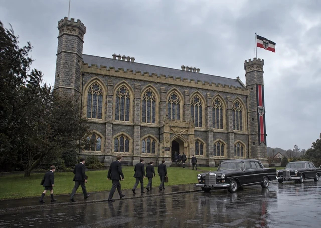 Fictional rainy 1964 exterior of Victoria College, with pupils crossing the lawn, period official cars and a restrained Reich flag on the historic school.